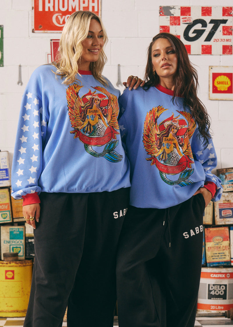 Two people wearing matching blue sweatshirts with a colorful design in a room with vintage signs and boxes.