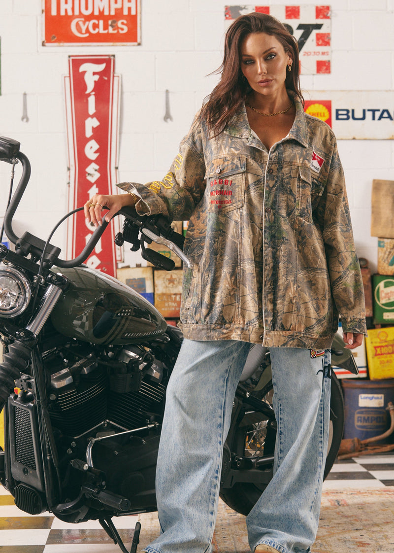 Woman in a camouflage jacket and jeans standing next to a motorcycle in a garage setting.
