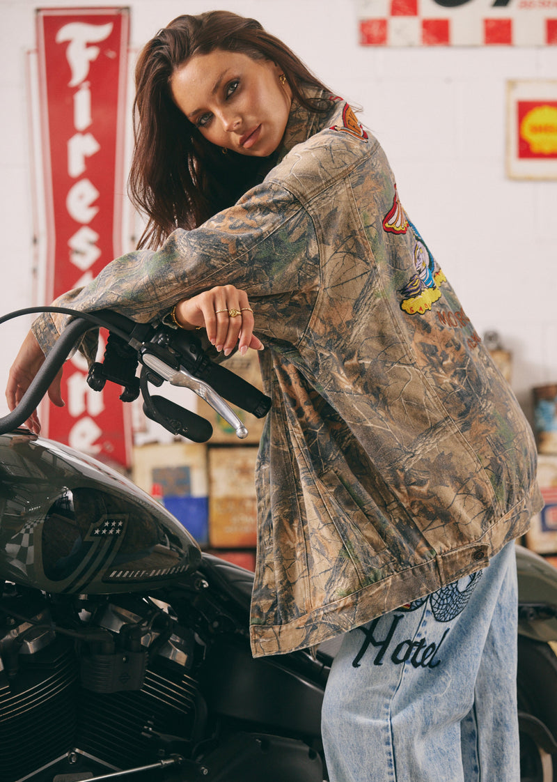 Person wearing a camouflage jacket with patches on a motorcycle in a garage setting