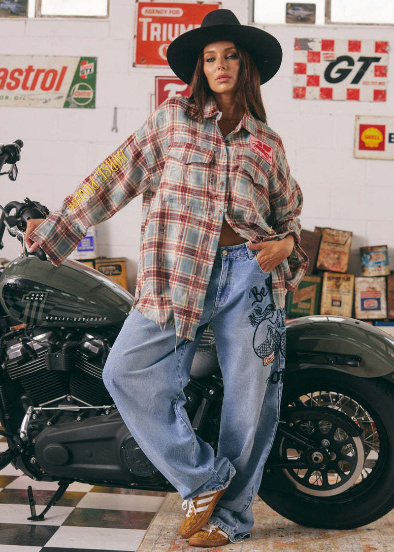 Person wearing a plaid shirt and jeans standing next to a motorcycle in a garage with vintage signs.