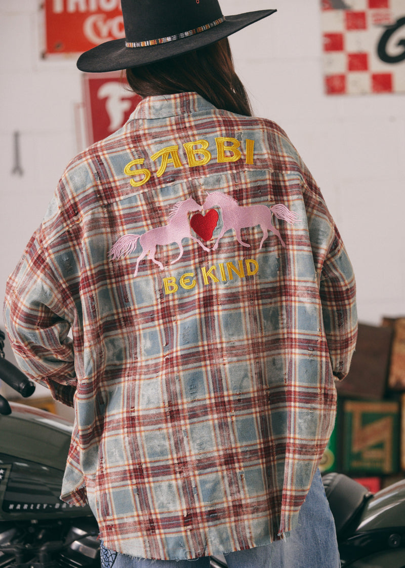 Person wearing a plaid shirt with 'SABBI' and 'BE KIND' text, standing in front of vintage signs.