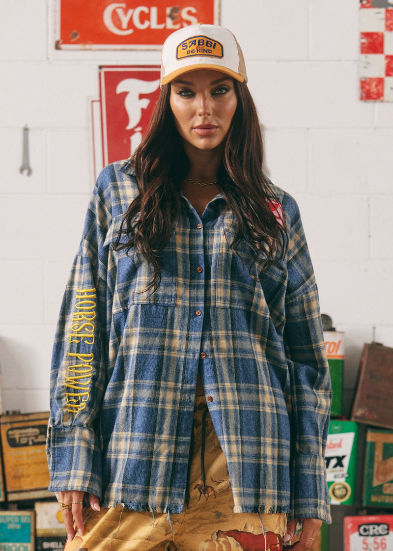 Woman wearing a plaid shirt and cap in a garage setting with vintage signs.