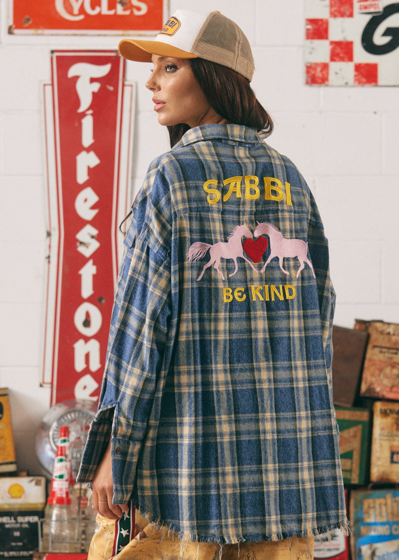 Person wearing a plaid shirt with 'SABBI BE KIND' text and horse design in a vintage setting.
